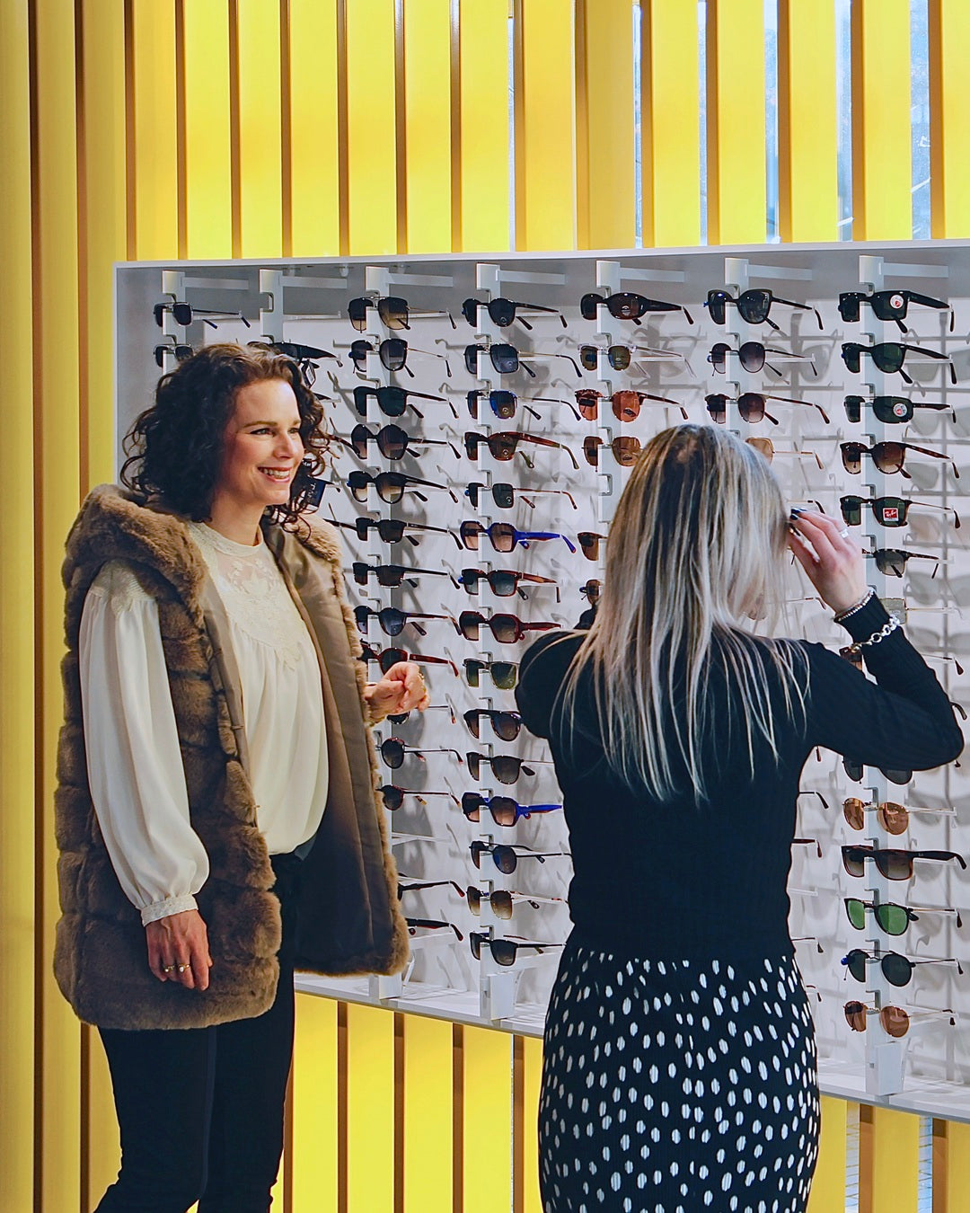 Two women looking at sunglasses on a eyewear display wall with a yellow background in a modern optical interior setting.